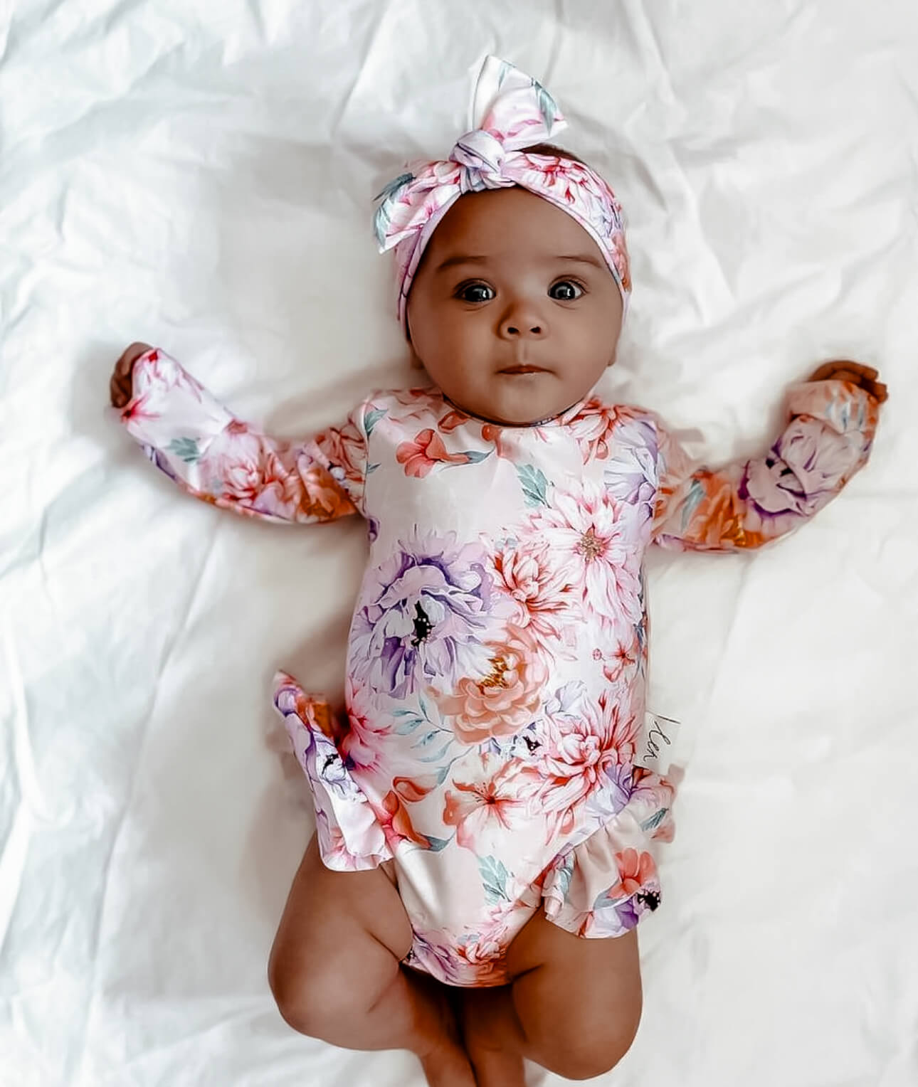 Baby girl wearing a floral sun safe swimsuit with nappy change snap onesie and headband on a white blanket