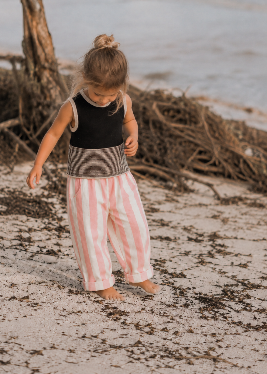 A toddler stands barefoot on a sandy beach wearing a sleeveless black top with a gray waist detail and pink-and-white vertically striped pants.