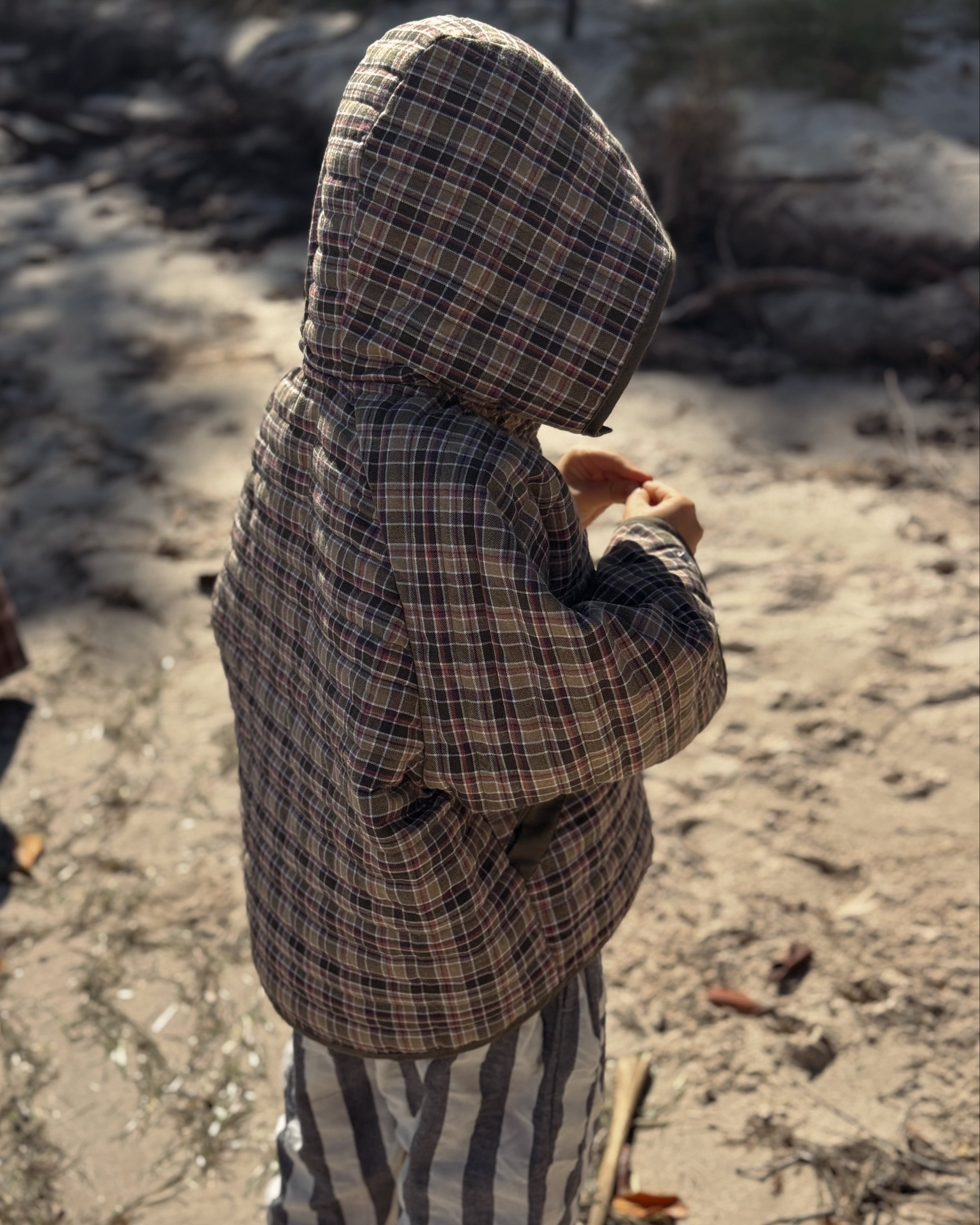 child wearing a plaid jacket and striped pants standing on a sandy or gravelly surface.