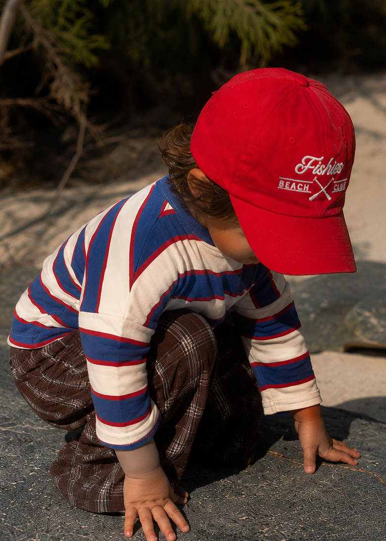 A young child crouching on a large rock outdoors, wearing a bright red baseball cap with white embroidered text reading 'Fight Like A BLACK' and a red, white, and blue striped long-sleeve shirt.