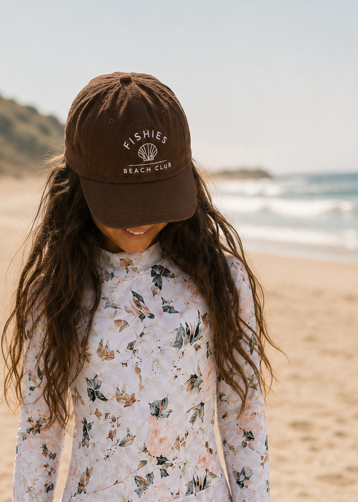 A young girl standing on a sandy beach wearing a brown baseball cap with 'FISHING BEACH CLUB' printed on it, and a long-sleeve white floral swim dress.