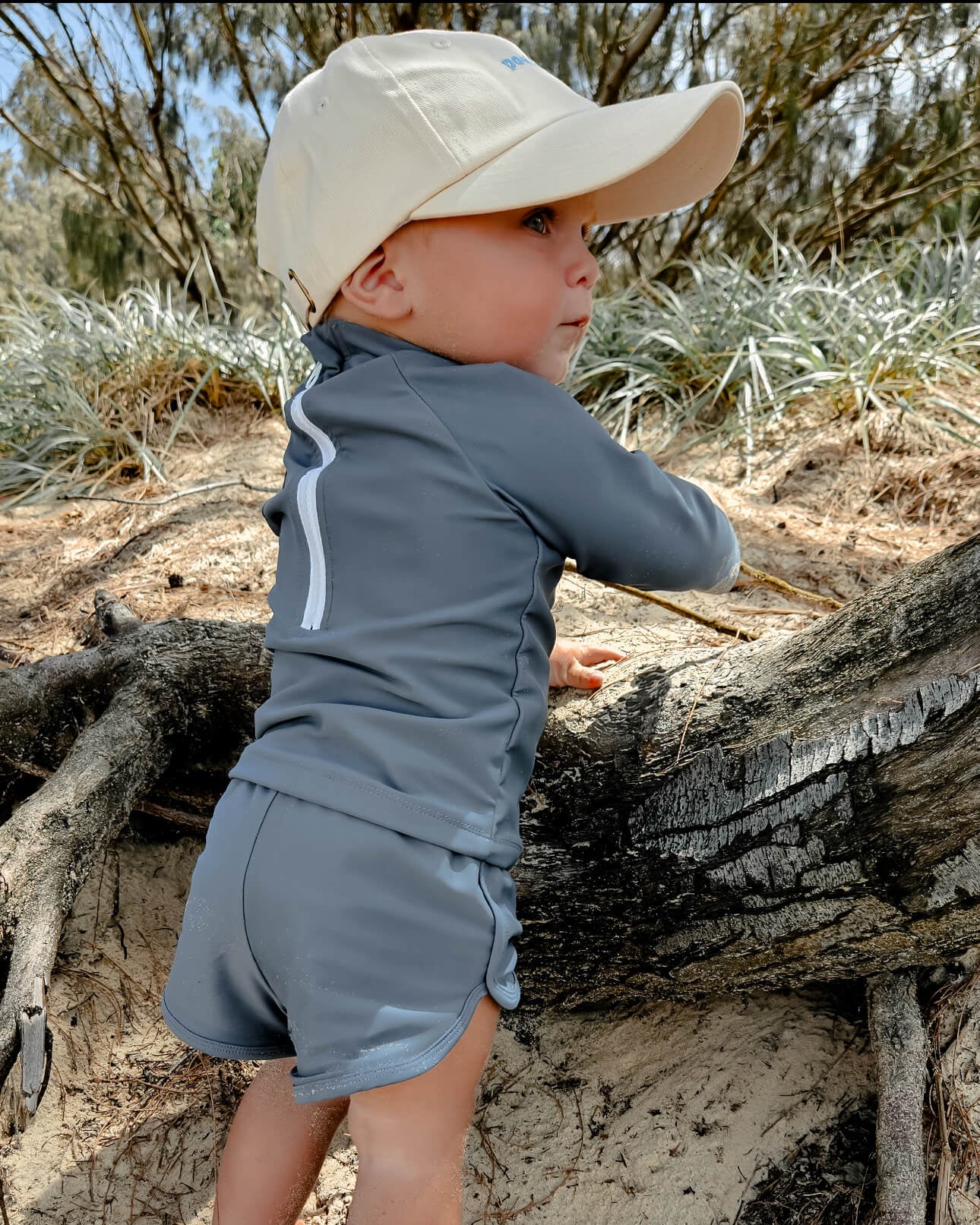 Child in sun safe long sleeve rash set and beige cap climbing a log in a natural setting