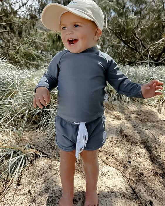 Child wearing a hat and navy rash set standing on a sandy surface with trees in the background