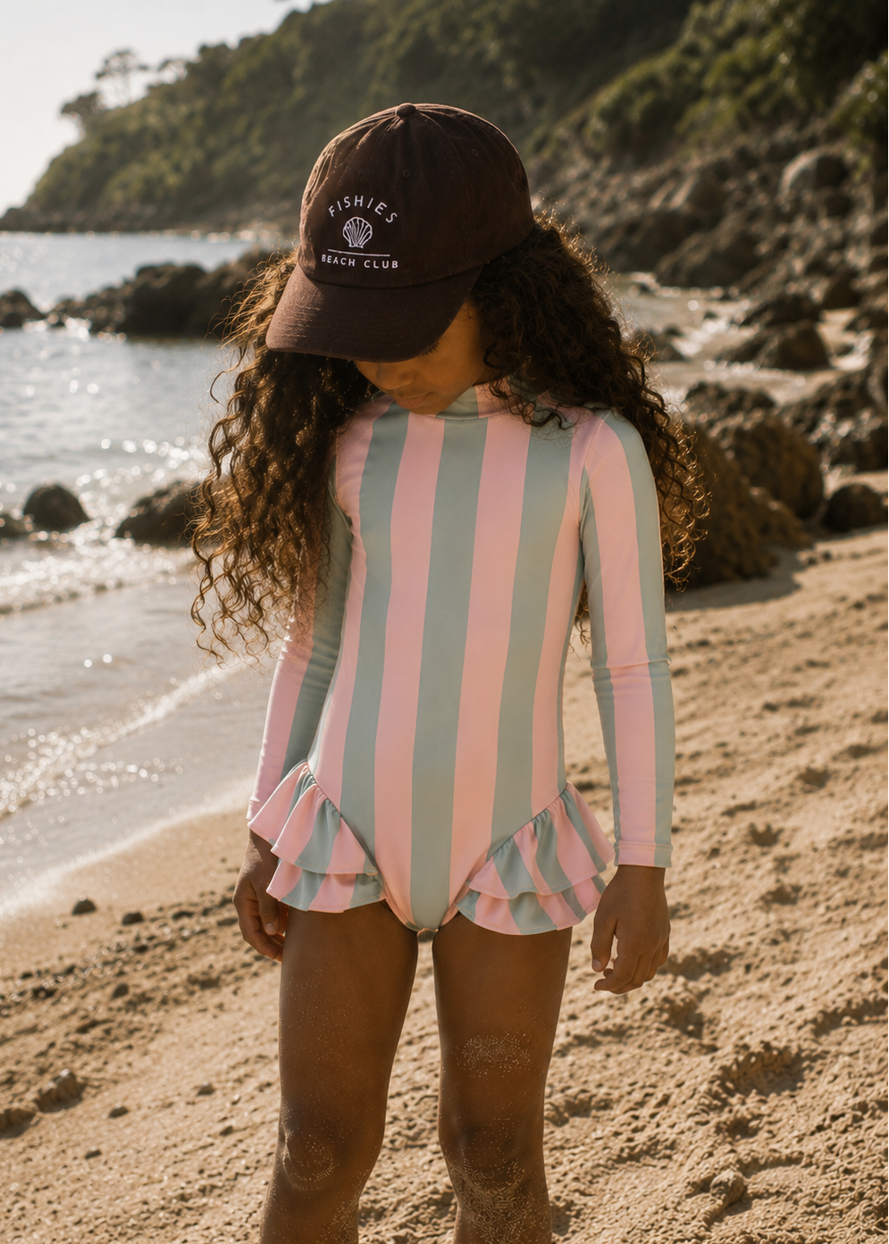A young girl standing on a sandy beach wearing a pink and mint green vertically striped long-sleeve swimsuit with a ruffled hem, a dark brown baseball cap with 'FISHIE'S WILD CLUB' printed on it, and curly hair.