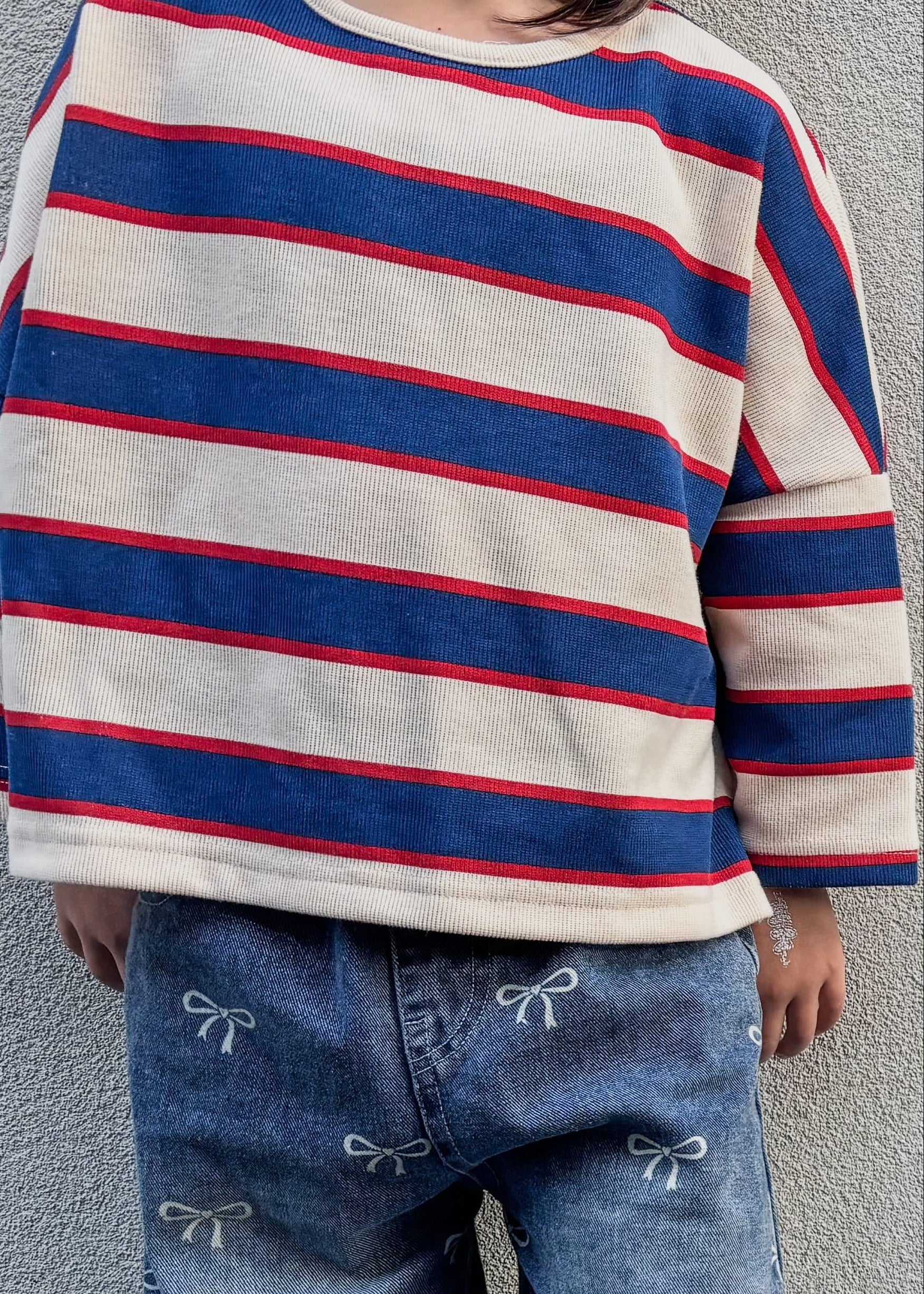 Child wearing a striped sweater and jeans standing against a plain wall.