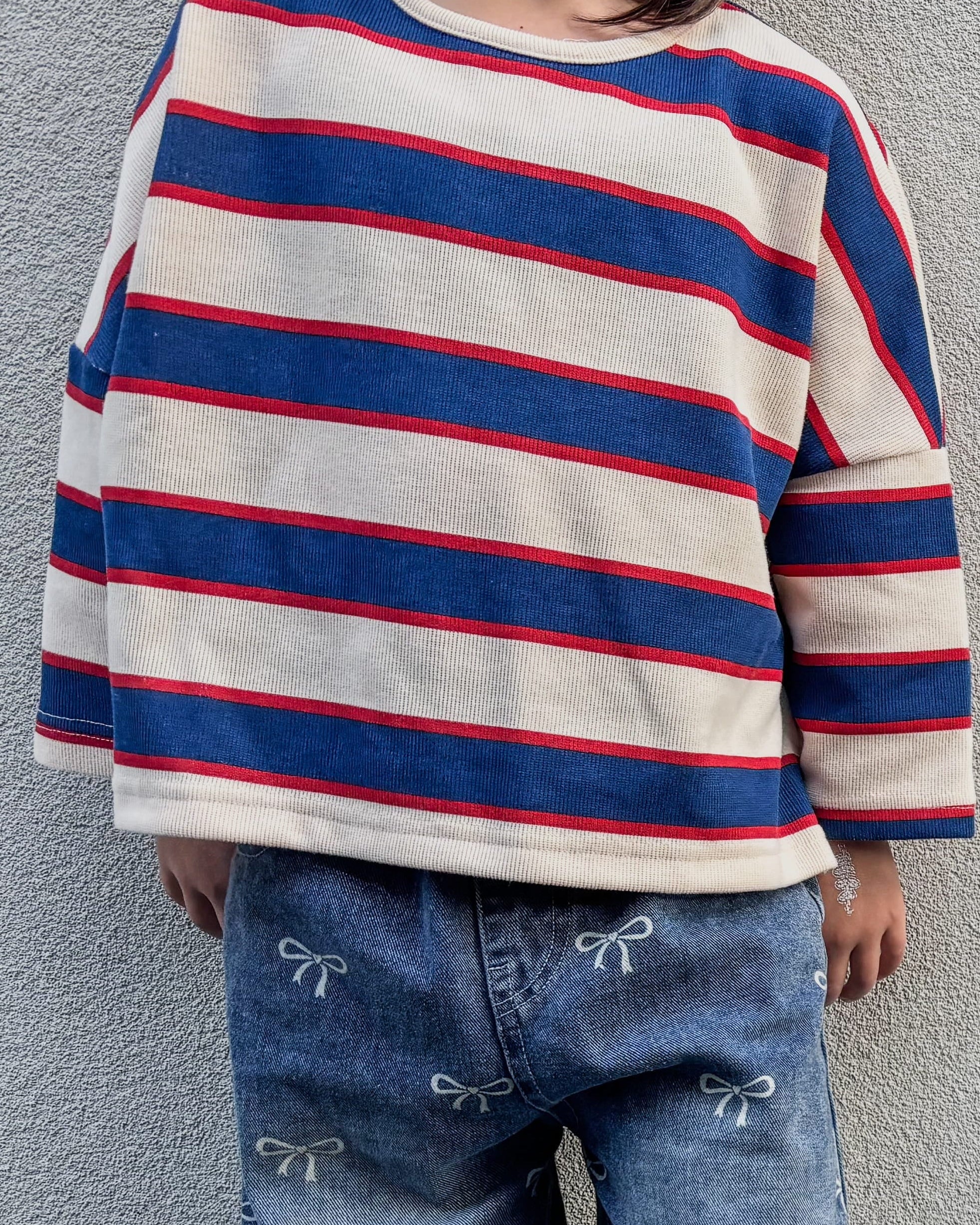 Child wearing a striped sweater and jeans standing against a plain wall.