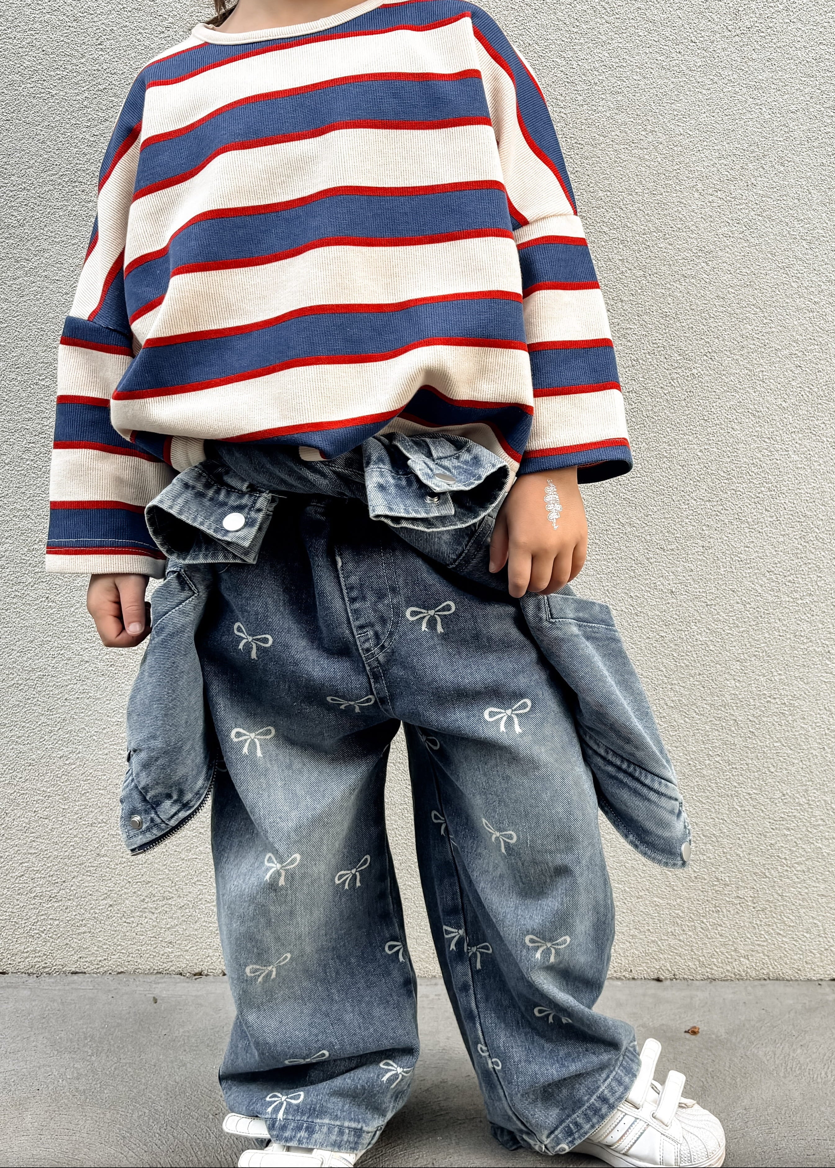 Child wearing a striped sweater and jeans standing against a plain wall.