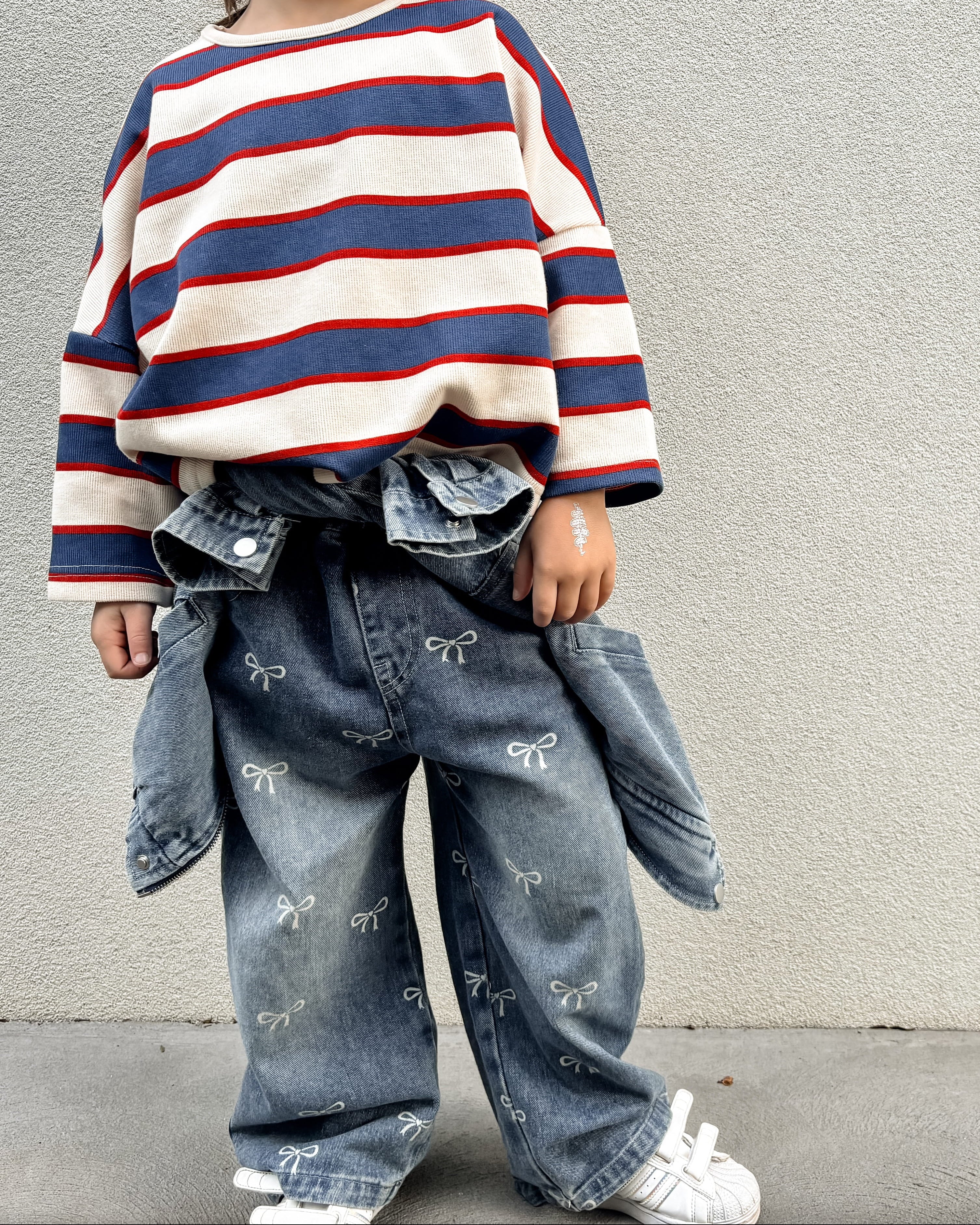 Child wearing a striped sweater and jeans standing against a plain wall.