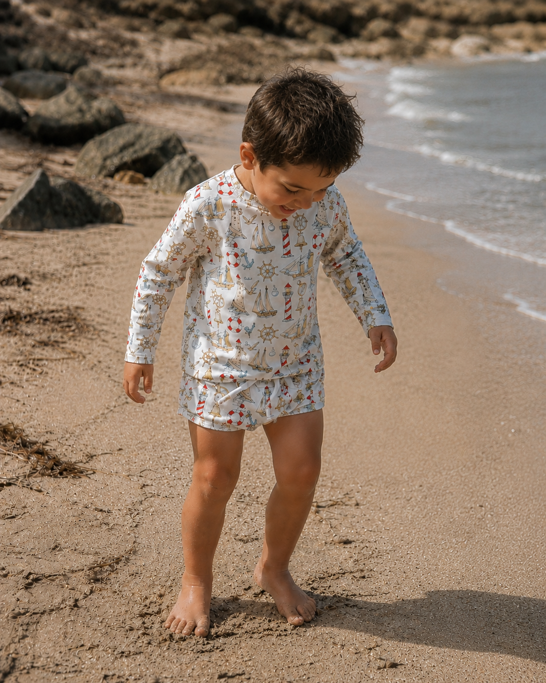 A young child walking barefoot on a sandy beach wearing a long-sleeve swim shirt and matching swim shorts with a white background and a red and green botanical pattern.