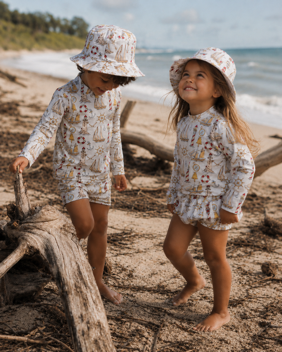 Two children in matching outfits and hats standing on a beach with driftwood.