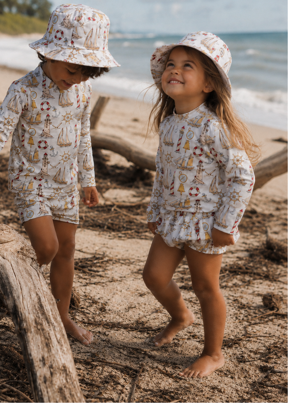 Two children in matching outfits and hats standing on a beach with driftwood.