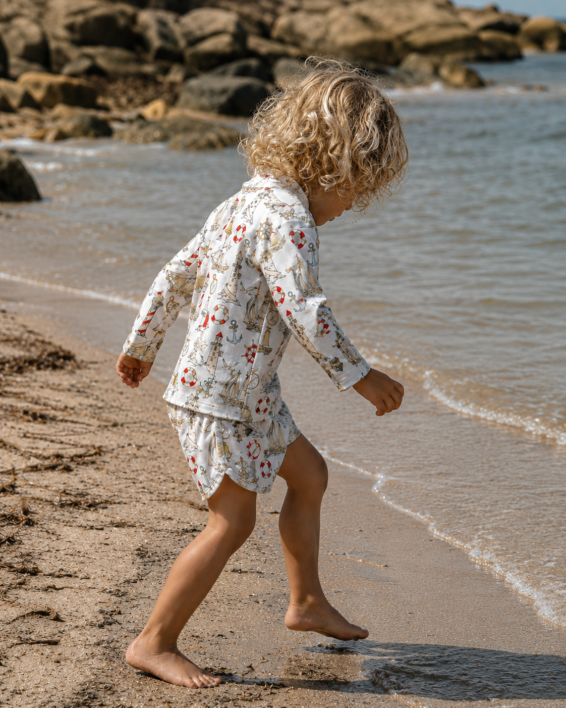 Child in a nautical rash set outfit walking on a beach with rocks and water in the background
