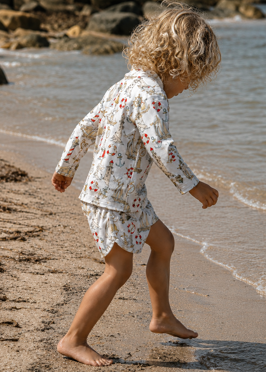 Child in a nautical rash set outfit walking on a beach with rocks and water in the background
