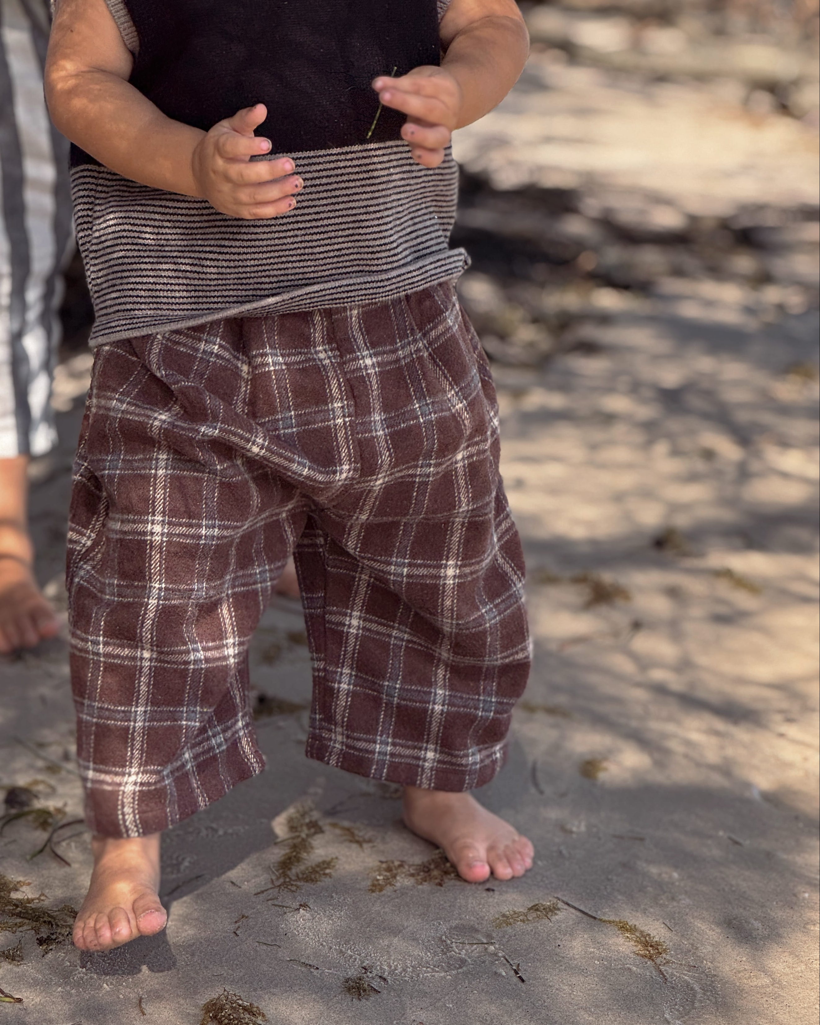 Child wearing a plaid outfit standing on a sandy beach with another person partially visible.