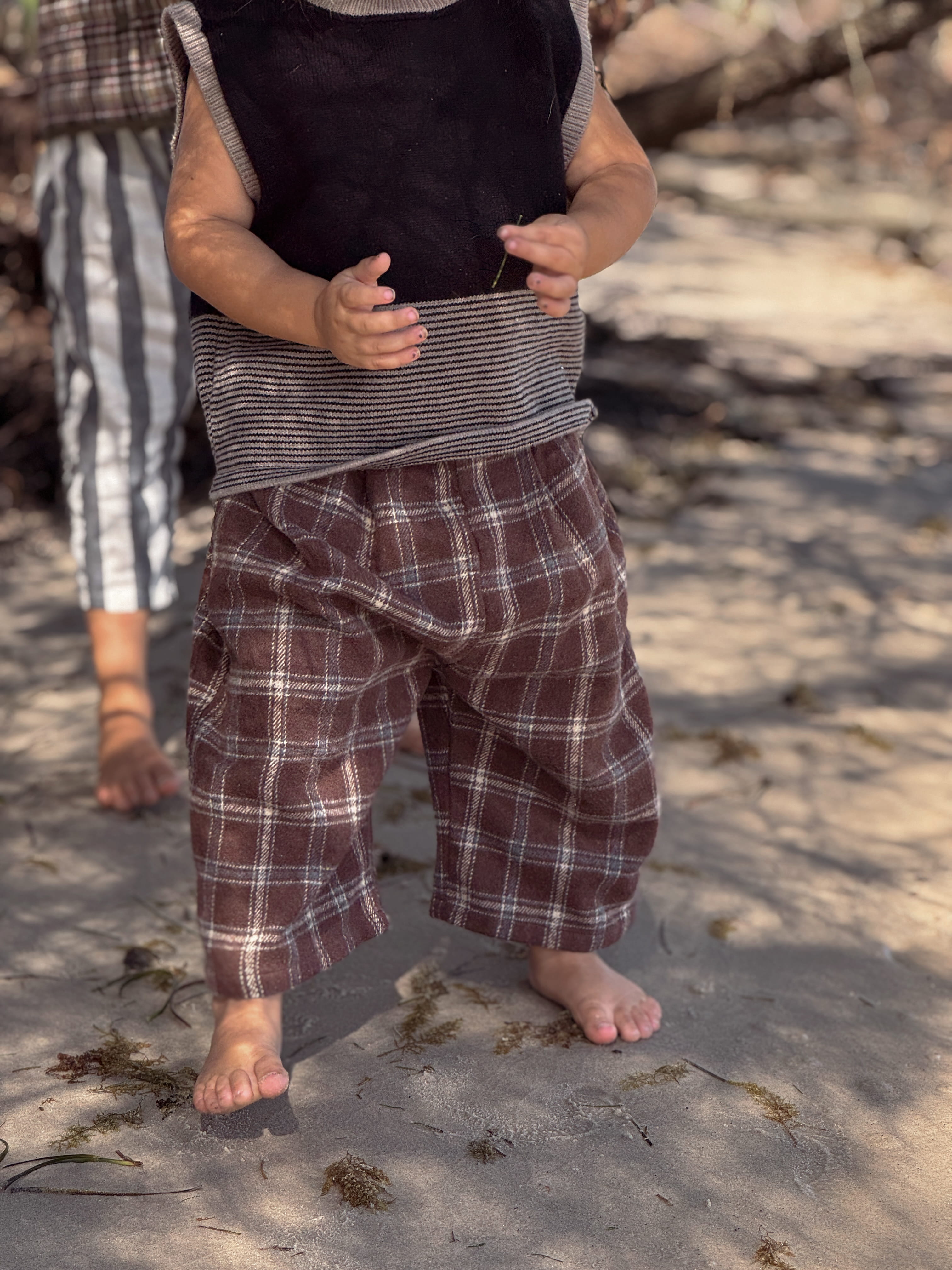 Child wearing a plaid outfit standing on a sandy beach with another person partially visible.