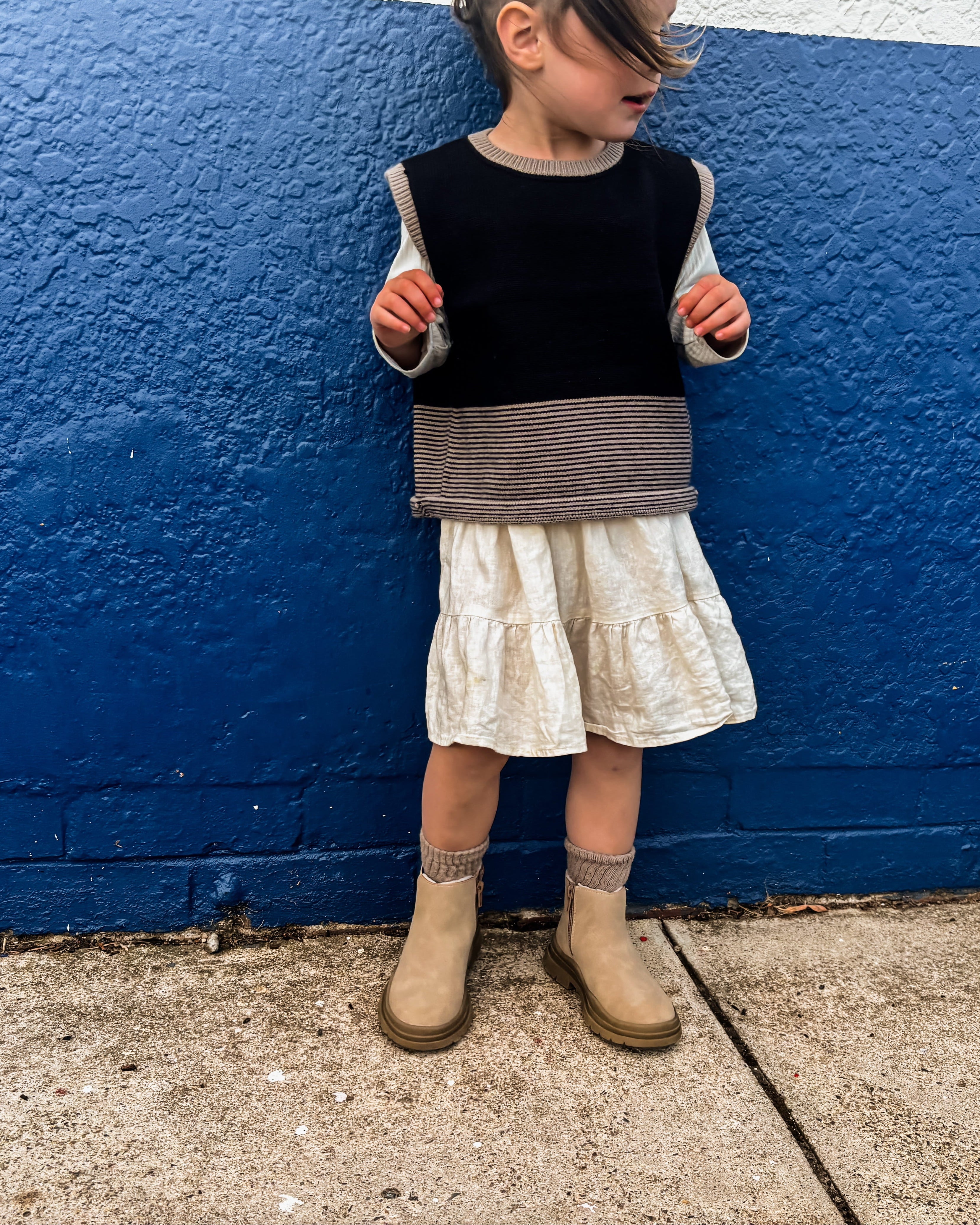 Child standing against a blue and white wall wearing a black sweater and white vest.