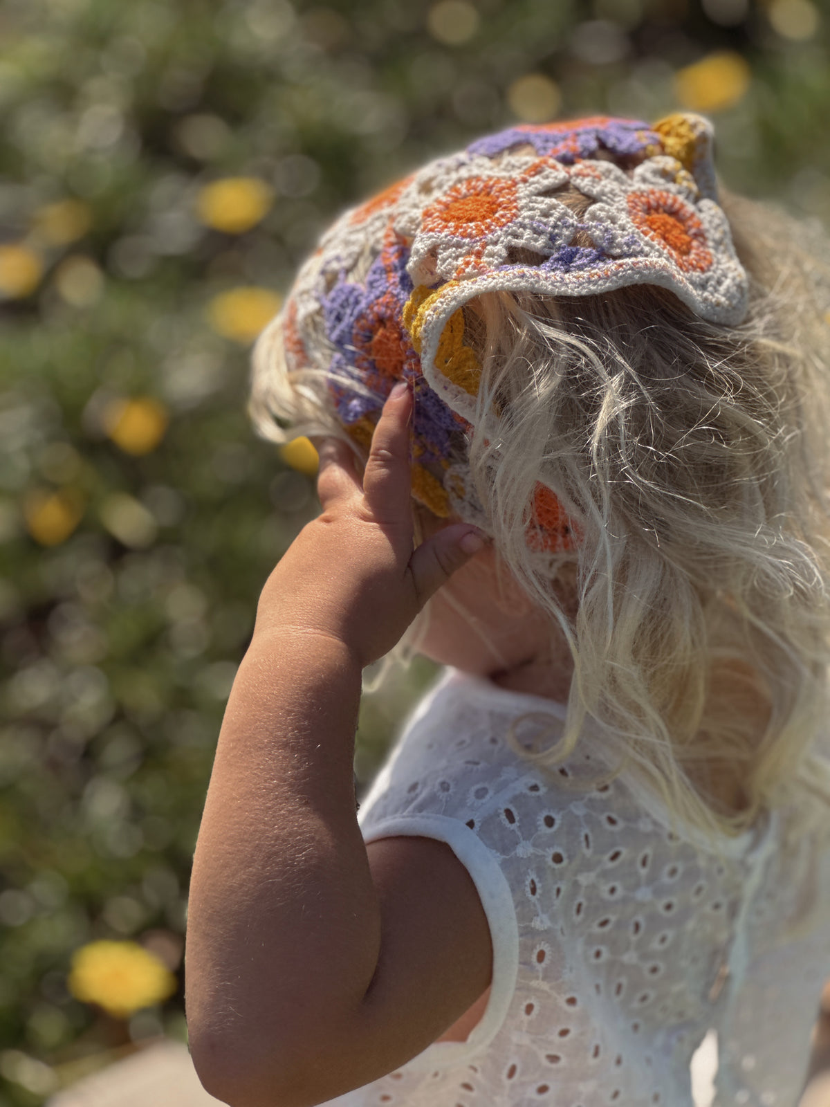 A young child wearing a colorful crocheted sun hat outdoors, with a white eyelet top and a blurred green garden background with yellow flowers.