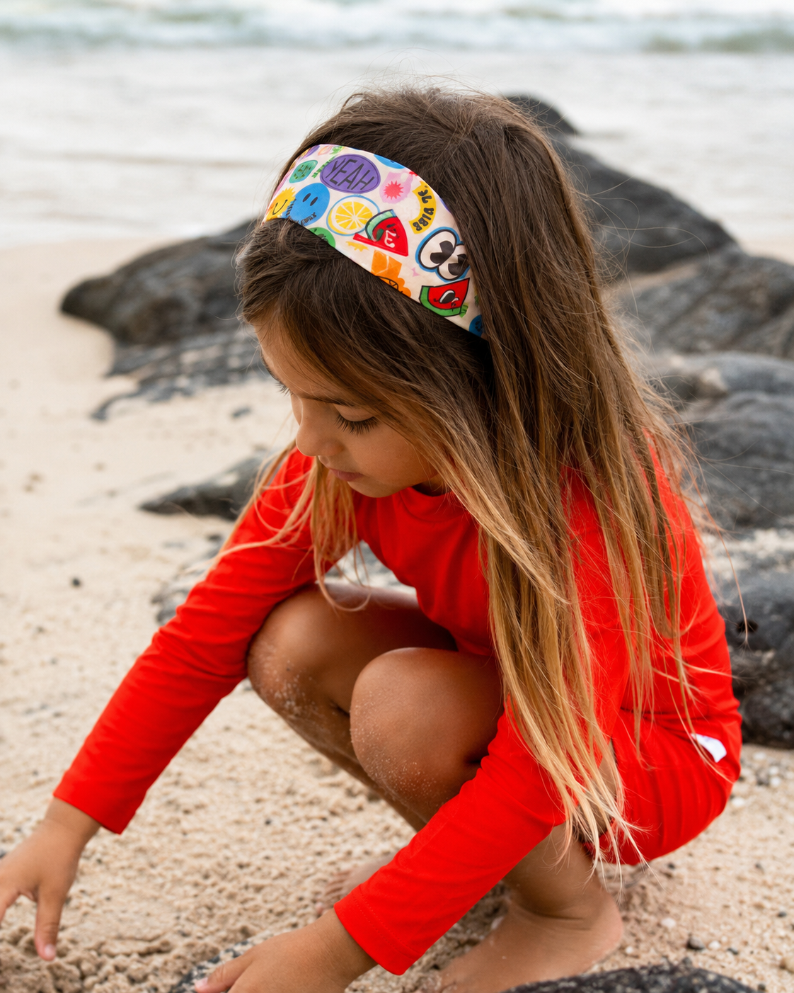 Child in a red rash suit and colorful headband playing on a beach.