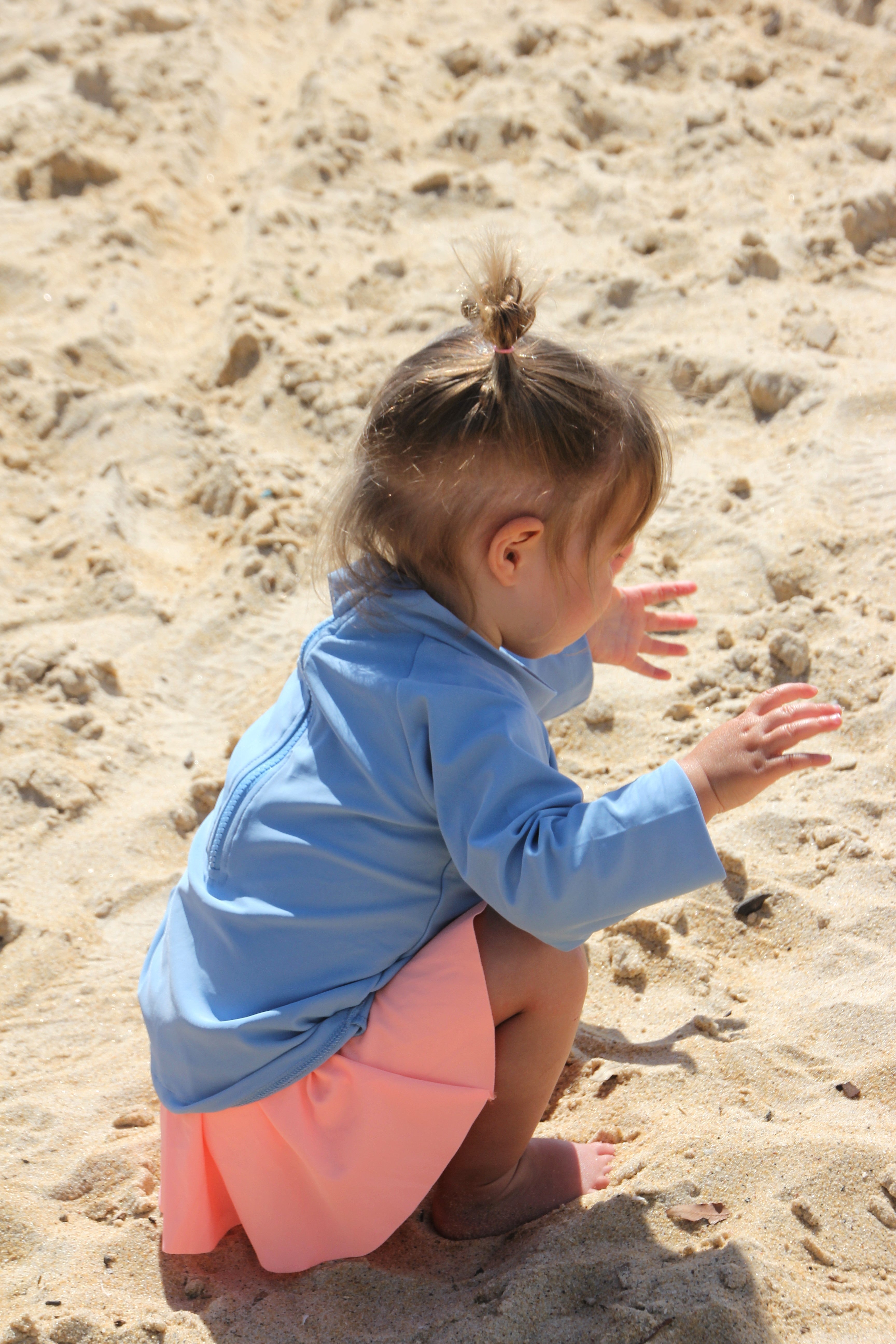 Young girl on the sand wearing a long sleeve blue rash guard top and an orange swim dress underneath