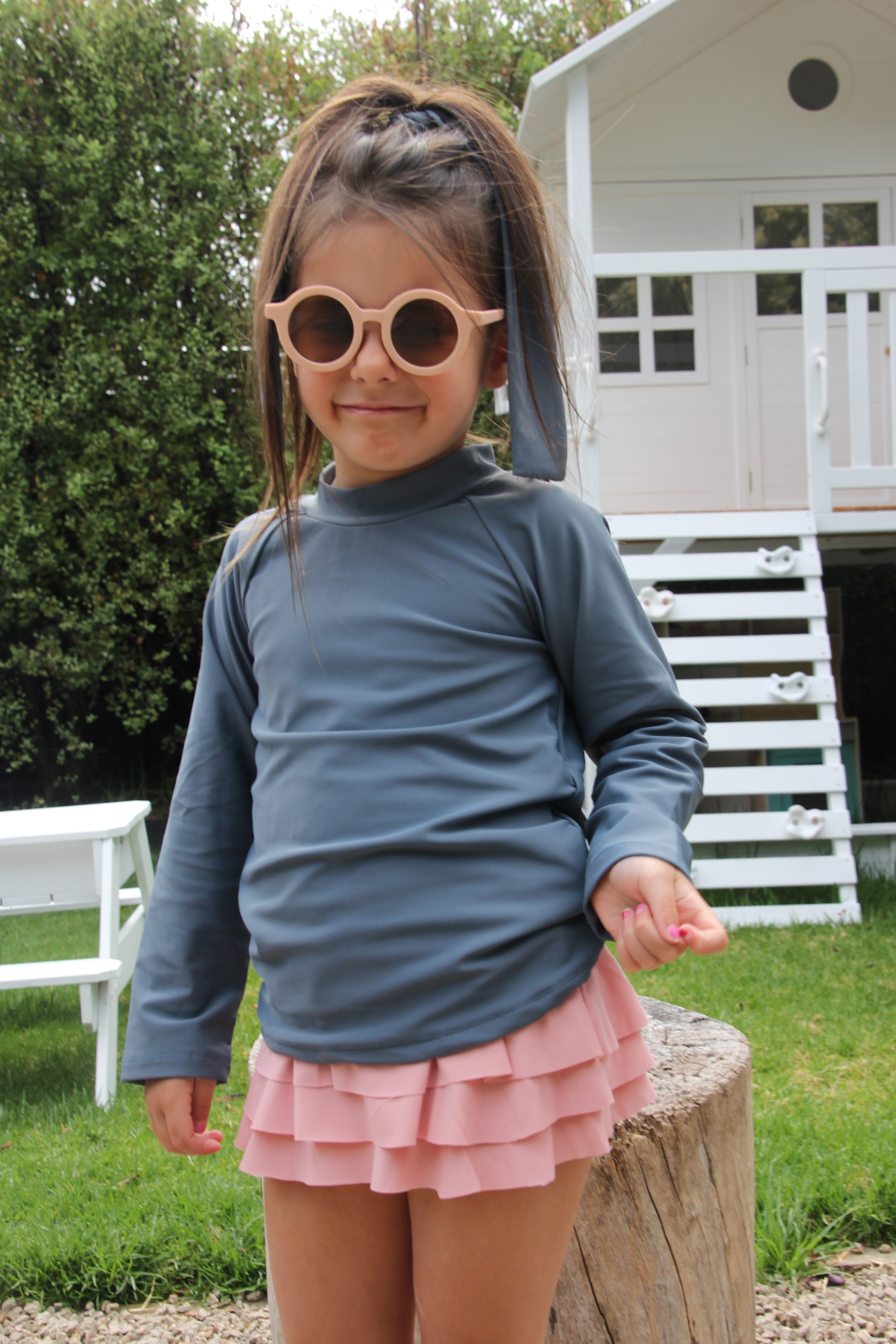 Girl wearing a blue rash guard vest and nappy changing friendly bloomer shorts with frills that are swim safe. She is standing in front of a cubby house