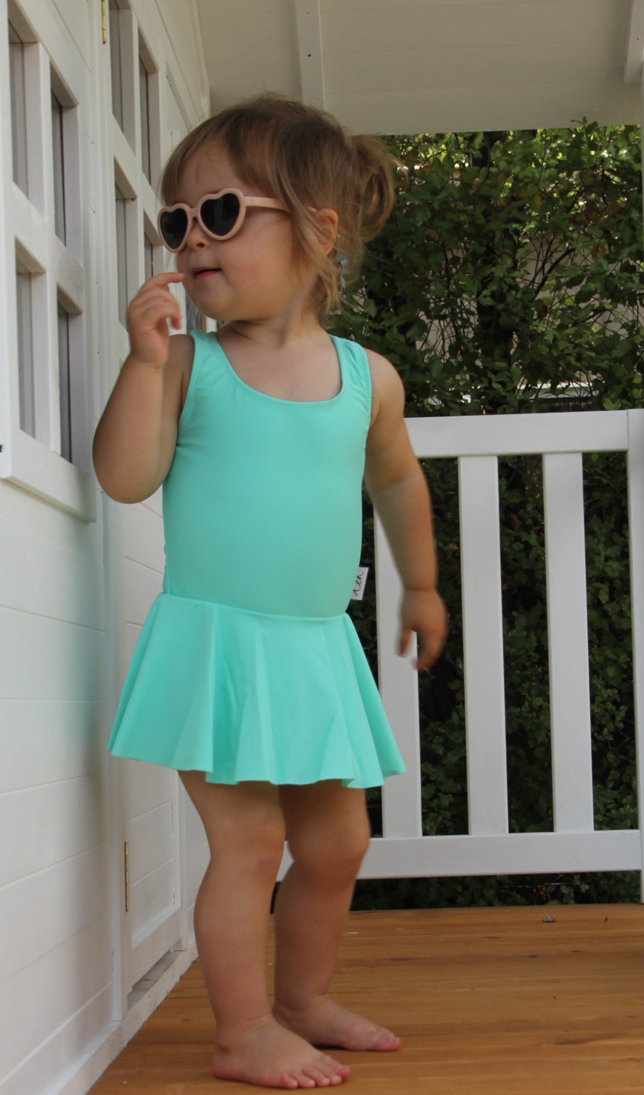 Baby toddler child wearing an aqua green swimsuit dress with snap closure at the bottom for nappy changes. Wearing heart sunglasses playing in a white cubby house