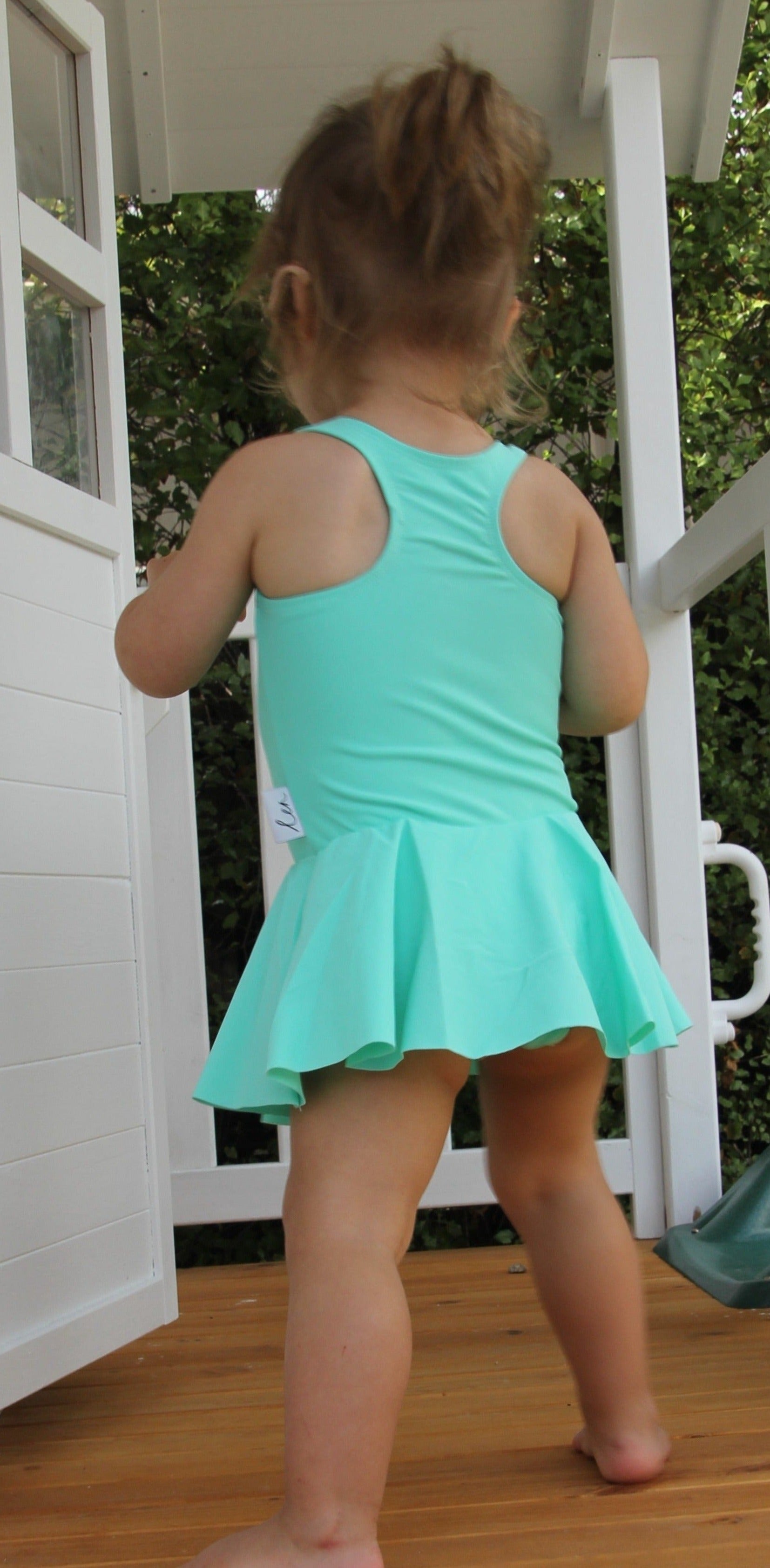 Baby toddler child wearing an aqua green swimsuit dress with snap closure at the bottom for nappy changes. They have their back to the image and are playing in a white cubby house
