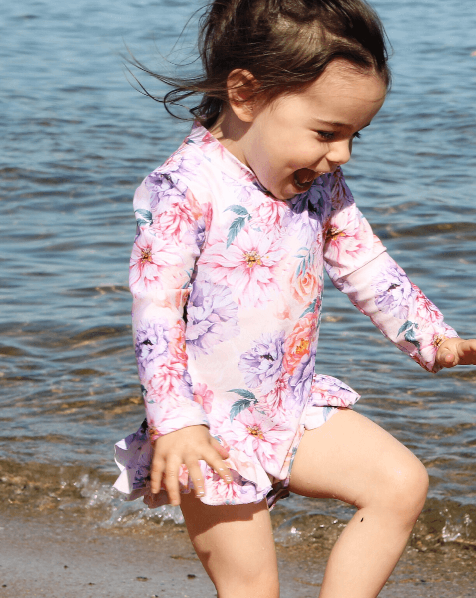 Baby, toddler and child swimsuit. The bathing suit is floral with orange and purple peonies and has nappy chainging snap closures at the bottom. It is on a little girl playing in the water on the sand at the beach. She is laughing